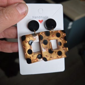 Op de afbeelding: Handgemaakte oorbellen met een zwart en goud design. De oorbellen hebben een vierkante vorm met een uitgesneden midden, zwarte stippen en gouden vlokken. De bovenkant van de oorbellen zijn zwarte cirkels. Ze zijn bevestigd aan een witte kaart met het woord "Handmade".