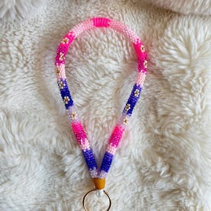 Pode incluir: Um cordão de contas em tons de rosa, roxo e branco, com detalhes dourados. O cordão tem um porta-chaves na parte inferior. As contas estão dispostas em um padrão de listras e flores. O cordão está sobre uma superfície branca e fofa.