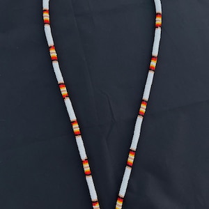 May include: A white beaded necklace with sections of red, orange, yellow, and black beads. The necklace is shown against a dark background. The beads are small and evenly spaced. The necklace is a traditional design.