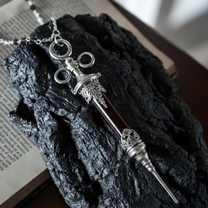 Pode incluir: Um colar com pingente prateado com um design em forma de seringa, detalhes ornamentados e um centro vermelho escuro. O colar é exibido em uma superfície escura e texturizada, evocando uma estética gótica ou steampunk.