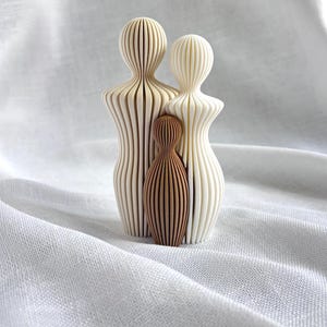 May include: A family of three wooden sculptures. The two larger figures are off-white, with a ribbed design. The smaller figure is brown and stands between the larger figures. The sculptures are on a white, textured surface.