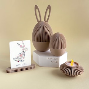 May include: An Easter-themed display featuring a bunny-eared egg, a smaller egg, a greeting card with a floral bunny design, and a candle holder with a flickering LED candle. The items are in shades of beige and brown.