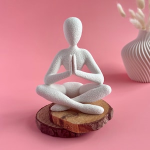 May include: A white textured yoga figurine in a seated pose with hands together, placed on two wooden slices. The figurine is set against a pink background, with a white vase and dried flowers in the background.