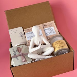 May include: A brown cardboard gift box filled with a white figurine in a seated yoga pose, tea packets, a candle labeled "White Lotus", and cards with botanical illustrations and text. The box is on a pink background.
