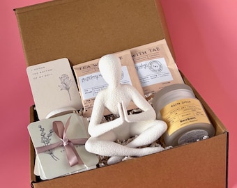 May include: A brown cardboard gift box containing a white yoga figurine, tea sachets, a "White Lotus" candle, and cards with floral designs and text. The box is set against a pink backdrop.