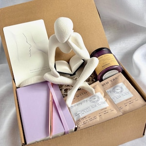 May include: A gift box filled with self-care items. Includes a white figurine, a lavender journal with a rose gold pen, tea sachets, a candle, and a card with a line drawing of a face. The box is brown.
