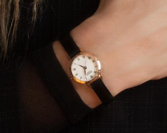 May include: A wristwatch with a round gold-tone case and a thin black strap. The watch face is white with black Roman numeral hour markers and black hands. The text "SOLEIL QUARTZ" is printed in black on the center of the white watch dial.