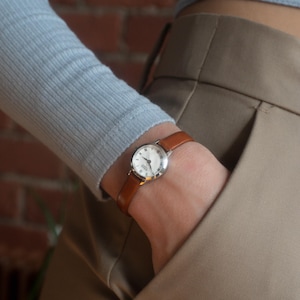 May include: A silver wristwatch with a white face and brown leather strap. The watch has Roman numeral hour markers. The watch is on a wrist, with the person wearing a light blue long-sleeved top and beige trousers.