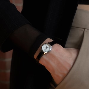 May include: A silver wristwatch with a black leather strap. The watch face is white with black Roman numerals and hands. The watch is worn on a wrist, with a black sleeve and beige trousers visible in the background.