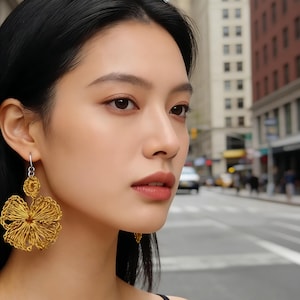 May include: A woman wearing gold-colored flower-shaped earrings. The earrings are made of wire and have a three-dimensional design. The woman is in an urban setting with buildings and a street in the background. The earrings are the focal point of the image.