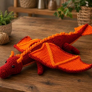 Puede incluir: Un juguete de dragón tejido a ganchillo hecho a mano con un cuerpo rojo oscuro, alas de color naranja brillante, una cresta dorsal naranja a juego y pequeños cuernos naranjas. La criatura tiene ojos de seguridad negros y una cola larga.