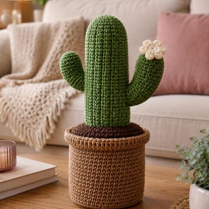 May include: A crocheted cactus in a tan pot with a dark brown soil base. The cactus is green with a textured surface, two side arms, and a small white flower attached to the right arm. The entire object is crafted from yarn with visible stitches.