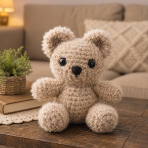 May include: A small, light beige crocheted teddy bear with a fuzzy texture, featuring two round dark brown eyes and a small black stitched nose. The bear is seated in an upright position with rounded ears, short arms, and legs extending forward.