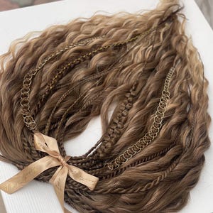 Könnte beinhalten: Eine Sammlung brauner und blonder Haarverlängerungen mit verschiedenen Texturen, darunter Zöpfe und wellige Abschnitte. Goldfarbene Kettenakzente sind in die Verlängerungen eingearbeitet. Ein beiges Band mit goldenem Text ist um die Basis gebunden.