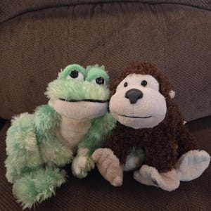 May include: Two plush toys: a green frog and a brown monkey. The frog has a light green body, white belly, and large black eyes. The monkey has brown fur, a white face, and tan hands and feet. Both are sitting.