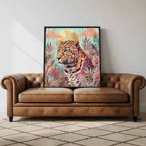 Puede incluir: Una impresión artística enmarcada que presenta un leopardo con un diseño floral colorido. El leopardo tiene un pelaje manchado y está situado sobre un fondo de luna dorada y follaje tropical. La obra de arte se exhibe sobre un sofá de cuero marrón.