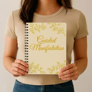May include: A spiral-bound notebook with the words "Guided Manifestation" in gold script. The notebook has a cream-colored cover with gold floral accents. The notebook is held by a person wearing a beige t-shirt and blue jeans.