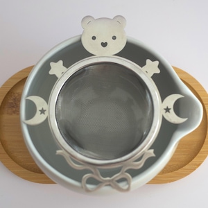 May include: A white ceramic tea mug with a metal mesh infuser insert. The infuser features a silver-toned rim decorated with a bear head, crescent moons, stars, and a bow. The mug rests on an oval-shaped light brown bamboo wooden tray.