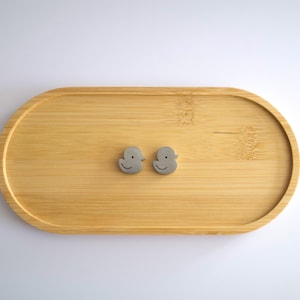May include: A pair of small, gray, duck-shaped stud earrings with simple etched facial features, positioned in the center of an oval-shaped, light-colored bamboo wooden tray with a smooth, natural grain finish and a raised edge.