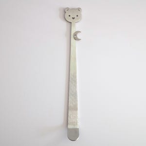 May include: A long, slender silver-toned metal bookmark featuring a flat bear-shaped head with a small heart-shaped nose and eyes, and a small crescent moon charm attached to the upper portion of the straight, flat stem.
