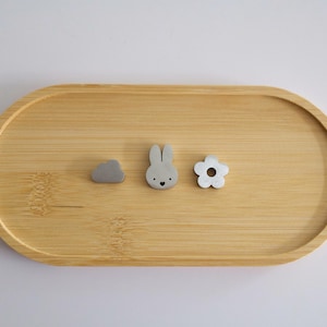 May include: An oval bamboo tray holding three small gray objects: a cloud shape, a rabbit head with two ears, and a five-petaled flower with a circular hole in the center. The tray has a smooth, light wood grain finish and raised edges.
