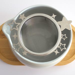 May include: A stainless steel tea strainer with a circular mesh basket and a wide rim featuring cut-out star and sparkle shapes. The strainer rests inside a white ceramic bowl, which is placed on an oval bamboo wooden tray.