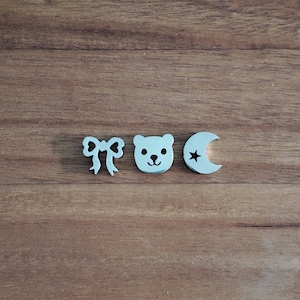 May include: Three light blue wooden cutouts: a bow, a bear face, and a crescent moon with a star. The objects are arranged on a brown wooden surface. The bear face has black eyes and a black nose.