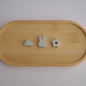 May include: A light brown bamboo tray with an oval shape. Three small, gray decorative objects are arranged on the tray: a cloud, a bunny, and a flower. The bunny has black eyes and a black nose. The tray is on a white surface.