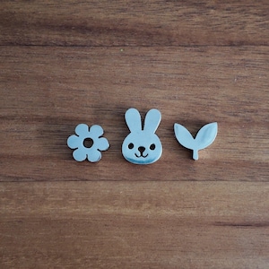 May include: Three small, silver-colored metal charms: a flower, a bunny, and a leaf. The bunny charm has a cute face with black dots for eyes and a small nose. The flower has six petals with a hole in the center. The leaf is a simple, curved shape.
