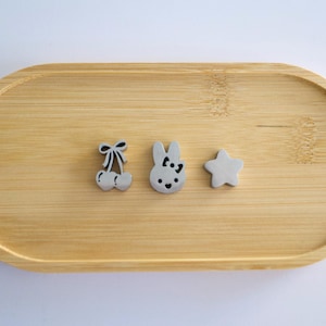 May include: Three small silver-toned metal charms shaped like a pair of cherries with a bow, a rabbit head with a bow, and a five-pointed star. The charms are arranged in a horizontal row on a smooth, light-colored wooden oval tray.