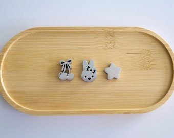 May include: Three small silver-toned metal charms shaped like a pair of cherries with a bow, a rabbit head with a bow, and a five-pointed star. The charms are arranged in a horizontal row on a smooth, light-colored wooden oval tray.