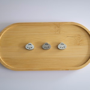 May include: Three small, light gray, rounded dumpling-shaped charms with black facial features, including eyes and mouths, arranged in a horizontal row on an oval-shaped, light brown bamboo wooden tray with a visible natural wood grain pattern.