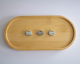 May include: Three small, light gray, rounded dumpling-shaped charms with black facial features, including eyes and mouths, arranged in a horizontal row on an oval-shaped, light brown bamboo wooden tray with a visible natural wood grain pattern.