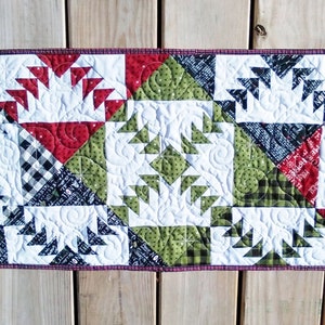 May include: A quilted table runner with a geometric pattern in red, green, black, and white. The runner is made of fabric squares with a quilted design.