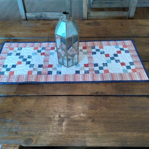 May include: A red, white, and blue quilted table runner with a geometric pattern. A clear glass lantern sits on top of the runner.