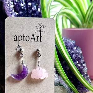 May include: A pair of earrings featuring a crescent moon made of amethyst and a cloud made of rose quartz. The earrings are displayed on a brown card with the text "aptoArt" and "aptoart.etsy.com".