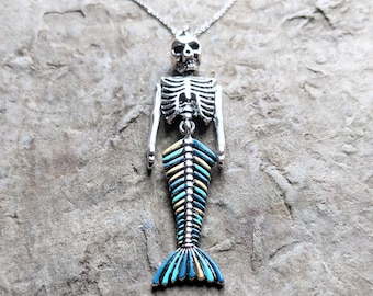 May include: A silver skeleton mermaid necklace with a blue and yellow tail. The necklace is on a gray textured background.