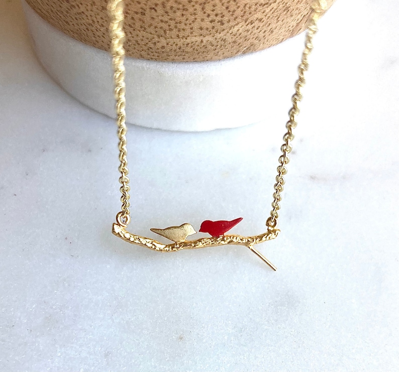 May include: A gold necklace with a delicate chain features two small birds perched on a gold twig. One bird is red and the other is white.
