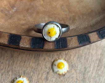May include: A silver ring with a white and yellow fried egg design.