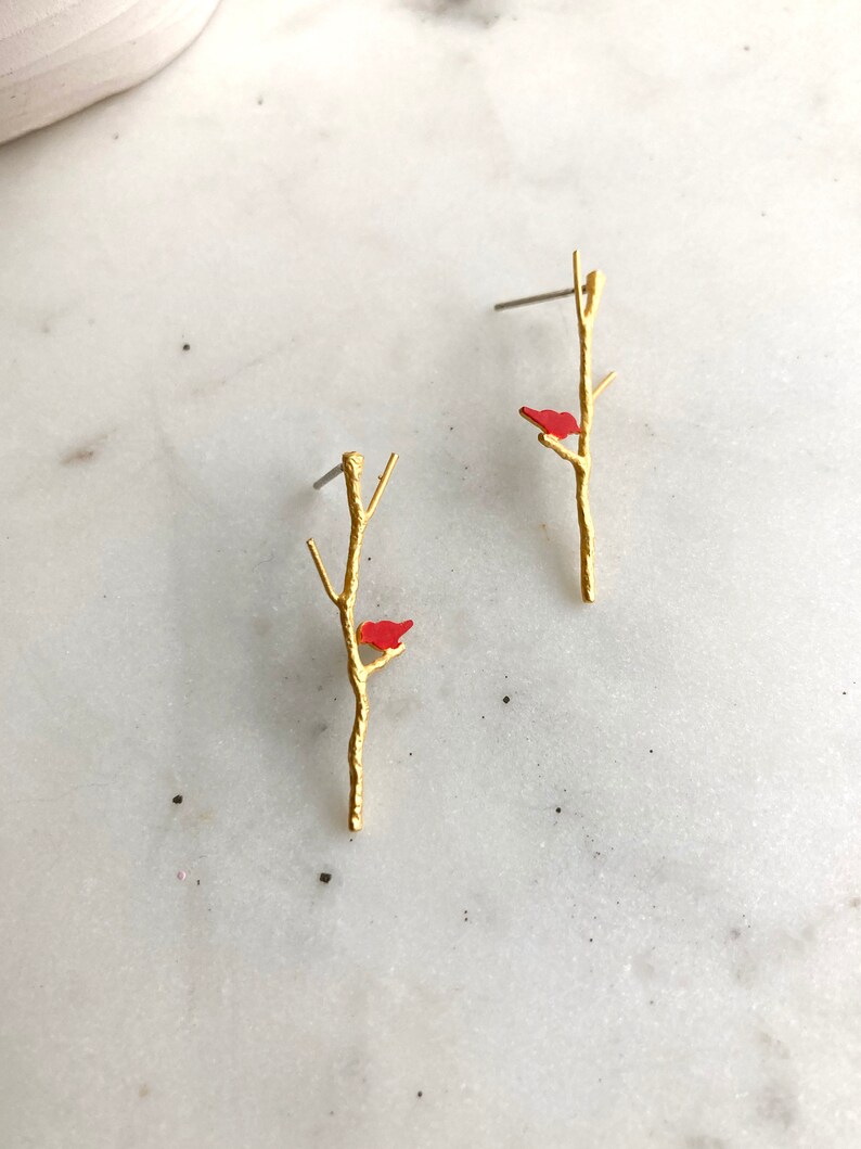 May include: A pair of gold earrings with a small red bird perched on a branch.