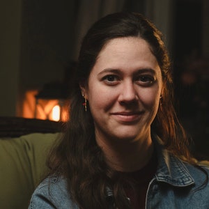 May include: A woman with long brown hair is wearing a denim jacket and smiling at the camera. She is sitting in a chair with a warm light behind her.
