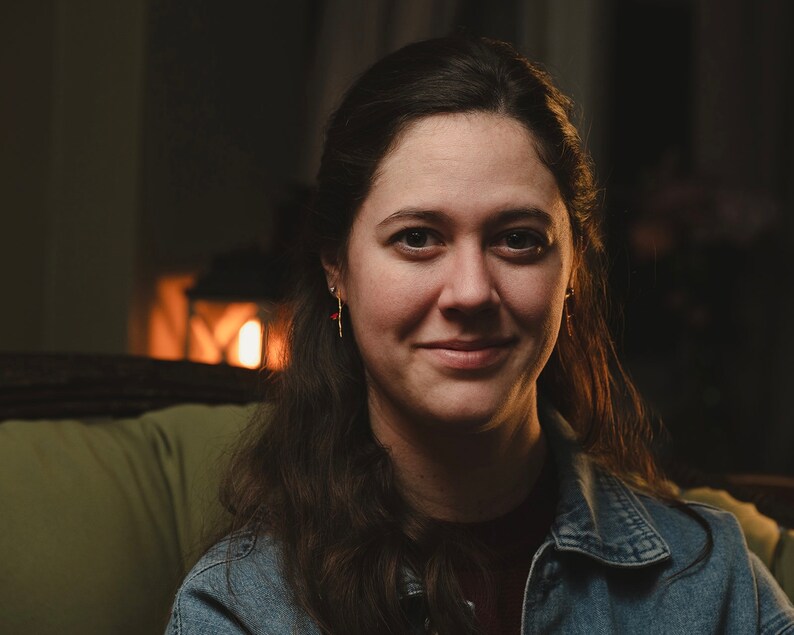 May include: A woman with long brown hair is wearing a denim jacket and smiling at the camera. She is sitting in a chair with a warm light behind her.
