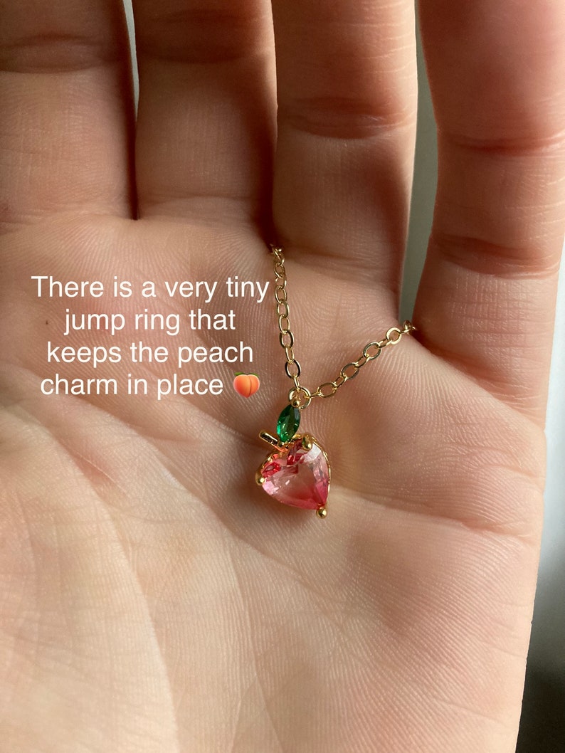 May include: A gold chain necklace with a peach-shaped charm. The charm is made of pink and clear glass and has a green leaf detail. There is a tiny jump ring that keeps the peach charm in place.