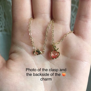 May include: Close-up of a gold chain necklace with a peach-coloured charm in the shape of a heart. The back of the charm is visible, showing a gold clasp.