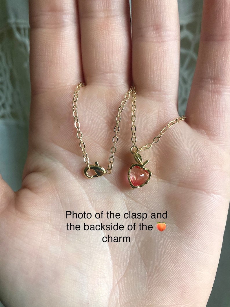 May include: Close-up of a gold chain necklace with a peach-coloured charm in the shape of a heart. The back of the charm is visible, showing a gold clasp.
