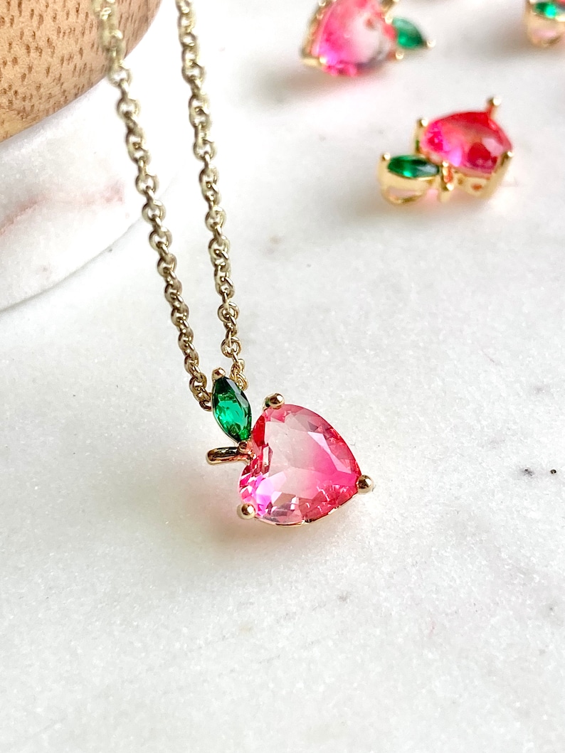 May include: A gold chain necklace with a pink heart-shaped pendant and green leaf accents. The pendant is made of clear glass with a pink hue.