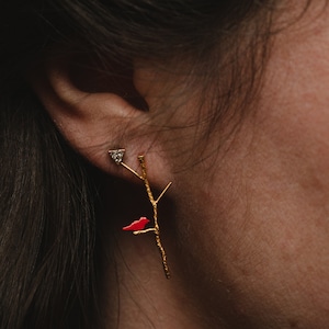 May include: Gold earrings with a red bird perched on a branch. The earrings feature a small diamond accent.