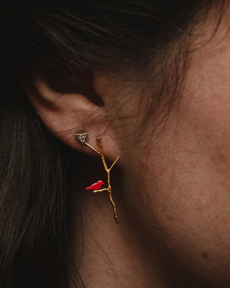 May include: Gold earrings with a red bird perched on a branch. The earrings feature a small diamond accent.