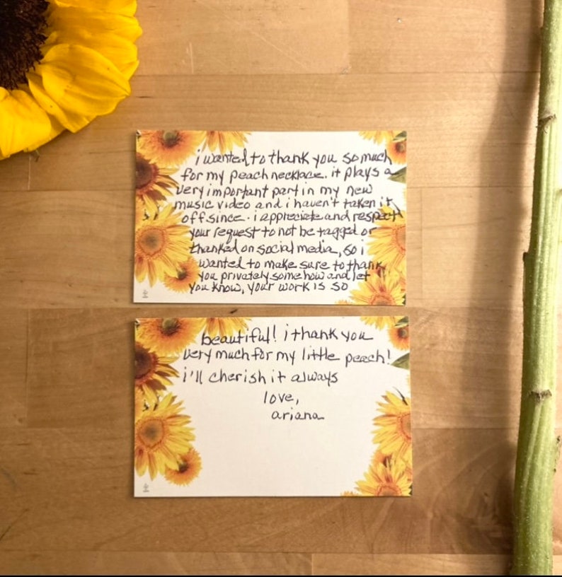 May include: Two handwritten thank you notes with sunflower designs. The first note says, "I wanted to thank you so much for my peach necklace. It plays a very important part in my new music video and I haven't taken it off since. I appreciate and respect your request to not be tagged or thanked on social media. So I wanted to make sure to thank you privately somehow and let you know, your work is so beautiful! I thank you very much for my little peach! I'll cherish it always. Love, Ariana."
