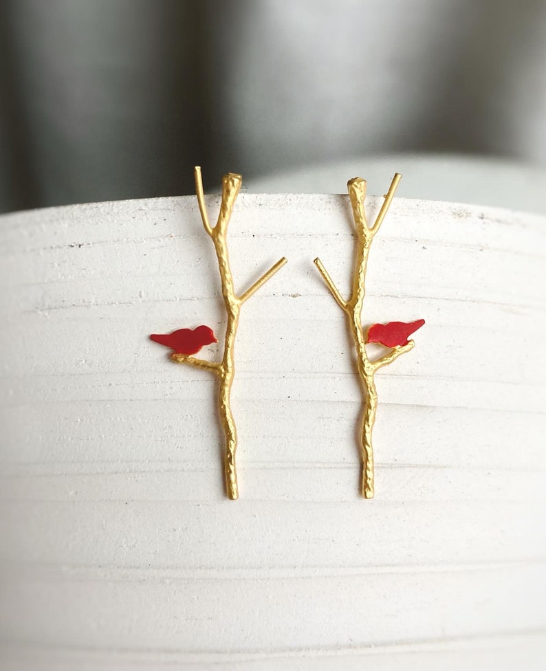 May include: A pair of gold earrings with a red bird perched on each branch. The branches are stylized to look like twigs.
